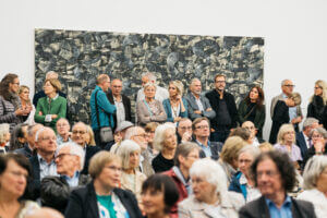 Besucher der Austellung stehen und Sitzen im Ausstellungsraum